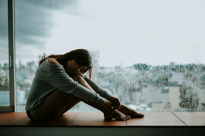 Person sitting on the floor hugging their knees by a rainy window